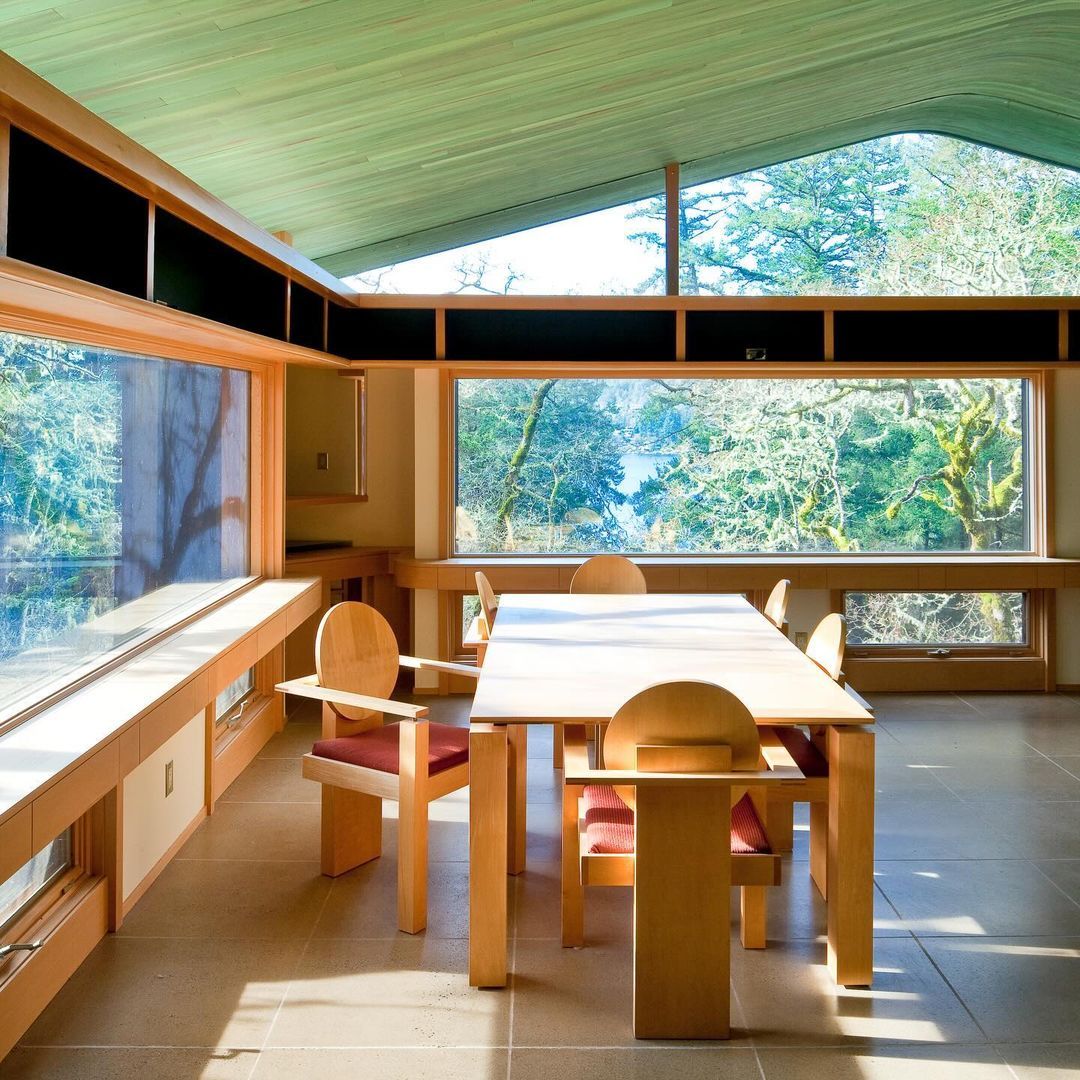 Dining area with a light-wood table looking out to the forest through a wall of windows beneath a sloped green ceiling.