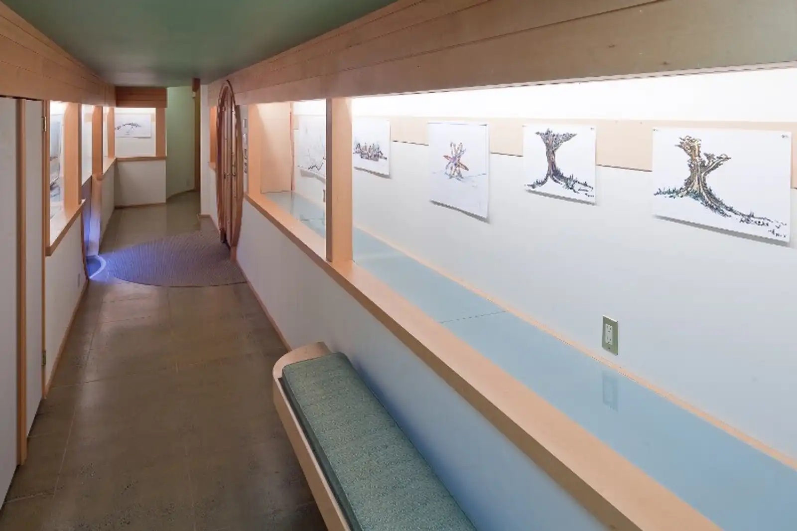 Long gallery hallway with artwork along the wall and a cushioned bench beneath a skylit band.